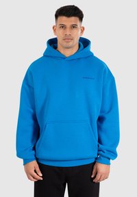 Felpa blu con tasca frontale e polsini a coste. Presenta una texture morbida e un logo nero discreto sul petto. Vestibilità semplice e rilassata.