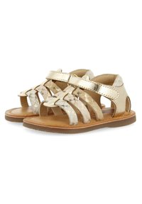 Gouden metallic leren sandalen met gevlochten banden, open zijkanten en een klittenbandsluiting. Bruine rubberen zool met een plat ontwerp.