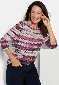 Langarmshirt in Pink, Weiß und Marineblau im Zickzackmuster. Runder Ausschnitt, weicher Stoff, kombiniert mit dunklen Jeans und einem marineblauen Gürtel.