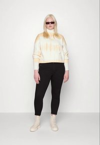 Sweatshirt com tie-dye branco e laranja, jeans skinny pretos e botas em camurça bege claro com atacadores. Modelo está de pé contra um fundo simples.