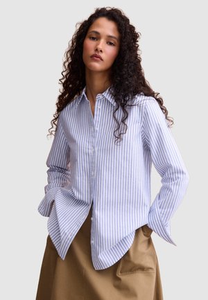 Donna con lunghi capelli ricci indossa una camicia bianca a righe azzurre con bottoni e una gonna beige, posando con le mani nelle tasche.