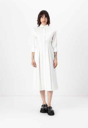 Robe blanche longueur genoux avec un col, manches 3/4 et une fermeture à boutons à l'avant. Présente une poche poitrine et une texture lisse.