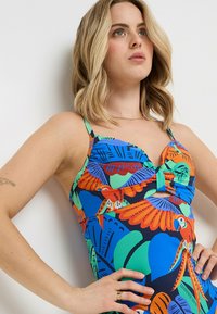 Maillot de bain à motifs multicolores avec des feuilles et des oiseaux bleus, verts, orange et rouges. Le matériau est lisse avec de fines bretelles réglables.