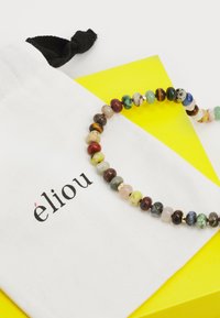 Κολιέ με χάντρες και πολύχρωμες πέτρες, με χρυσές λεπτομέρειες, τοποθετημένο σε ένα λευκό υφασμάτινο σακουλάκι με το λογότυπο "éliou", δίπλα σε μια φωτεινή κίτρινη τριγωνική κάρτα.