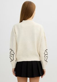 Suéter color crema de manga larga, con bordados florales en negro en la manga izquierda, y puños y dobladillo acanalados. Combinado con una falda negra con pliegues.
