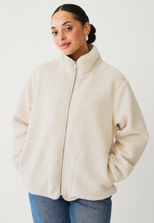 Cream fleece jacket with high collar, full zipper, and front pockets. Soft texture, relaxed fit, and simple design. Worn over jeans.