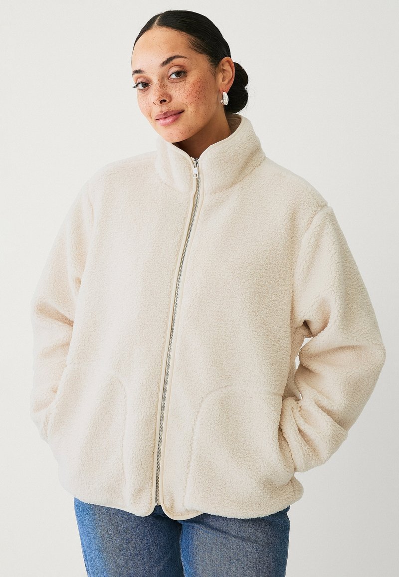 Cremfarbene Fleecejacke mit hohem Kragen, durchgehendem Reißverschluss und vorderen Taschen. Weiche Textur, lässige Passform und schlichtes Design. Über Jeans getragen.