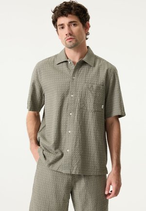 Camisa de manga corta con botones en tela texturizada verde claro, con un pequeño bolsillo en el pecho y dobladillo recto. Pantalones cortos a juego visibles.