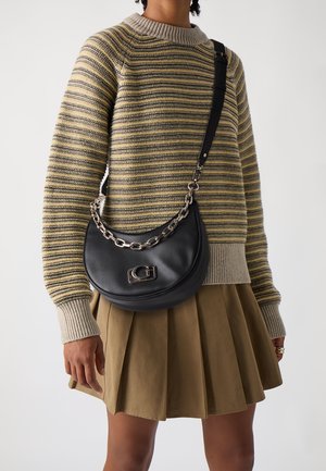 Sac à bandoulière en cuir noir avec une forme arrondie, une sangle en chaîne et des accessoires métalliques. Porté sur un pull rayé et une jupe plissée olive.