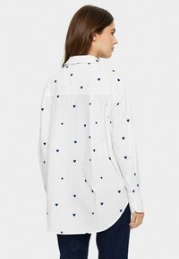White shirt featuring small navy heart patterns, button-up design, long sleeves, and a curved hem. Material appears lightweight and smooth.