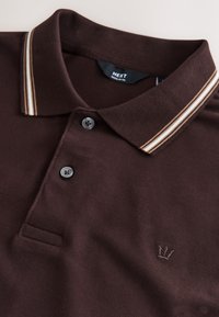 Polo marron foncé avec deux boutons, col rayé blanc et beige, et petit logo couronne brodé sur la poitrine.