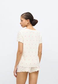 Σετ ύπνου με κοντά μανίκια σε κρεμ απόχρωση με ροζ μοτίβα καρδιάς. Τα κυματιστά άκρα στο πάνω μέρος και το σορτς προσθέτουν υφή. Μαλακό ριμπ ύφασμα.