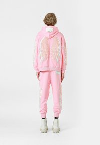 Conjunto de sudadera y pantalones de chándal rosa con grandes diseños de alas bordadas en la parte posterior, con puños acanalados y botas blancas resistentes.