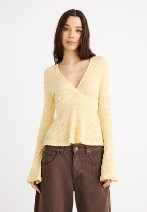 Ung kvinne med langt mørkt hår iført en strukturert, lys gul V-hals langermet topp og løse brune bukser mot hvit bakgrunn.