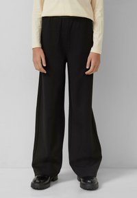 Zwarte broek met wijde pijpen gemaakt van zachte stof, voorzien van een elastische tailleband en een naadloos ontwerp. Gecombineerd met chunky zwarte schoenen.