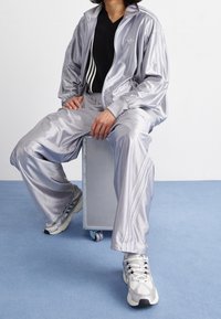 Osoba oblečená ve stříbrné lesklé teplákové soupravě a bílých teniskách sedící na malé bedně na kolečkách na modré podlaze.