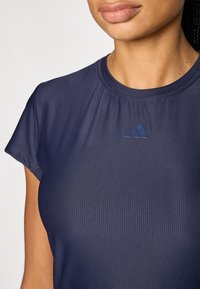 Camiseta deportiva de manga corta en azul marino con cuello redondo, textura acanalada y un pequeño logo azul de Adidas en el pecho.