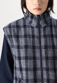 Bezgala veste zils un pelēks pārbaudījuma raksts. Izgatavota no teksturēta auduma ar augstu apkakli un pilnu priekšējo rāvējslēdzēju. Nav redzamu kabatu.