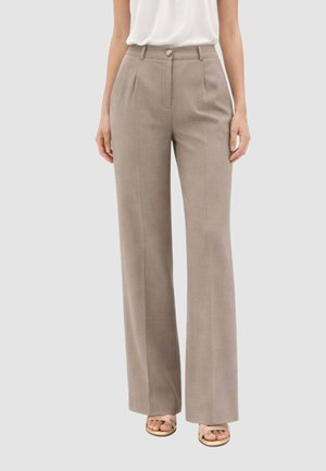 Donna che indossa pantaloni taupe a gamba larga a vita alta con pieghe frontali e una camicetta bianca senza maniche infilata dentro, in piedi davanti a uno sfondo neutro.