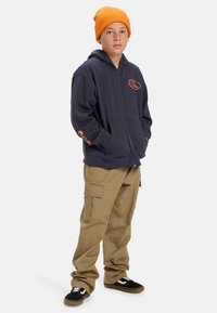Navy zip-up hoodie with orange logo, front pockets, paired with beige cargo pants and black sneakers with white accents, topped with an orange beanie.