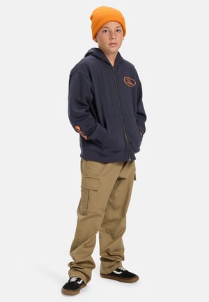 Marineblauwe rits hoodie met oranje logo, voorzakken, gecombineerd met beige cargobroek en zwarte sneakers met witte accenten, afgewerkt met een oranje muts.