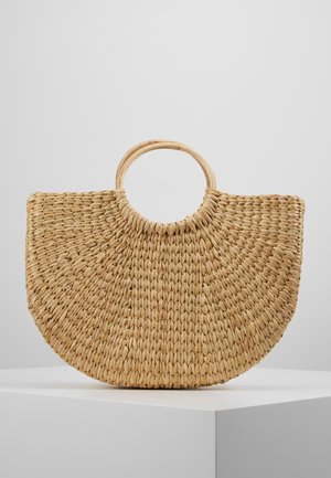 Gevlochten strohandtas met halfcirkelvorm en ronde houten handgrepen, tentoongesteld op een wit voetstuk tegen een effen achtergrond.