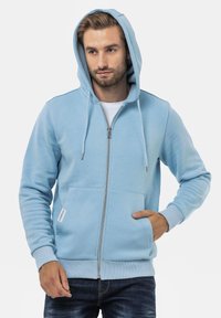 Hoodie bleu clair avec fermeture éclair à l'avant, capuche avec cordon de serrage et deux poches avant. Il présente des poignets et un ourlet côtelés, avec une texture de tissu lisse.