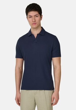 Mężczyzna-model ubrany w granatową koszulkę polo z krótkim rękawem i beżowe spodnie, stojący na tle jednolitego jasnoszarego tła.