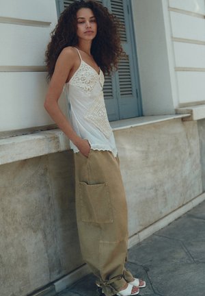 Femme aux cheveux bouclés portant un caraco en dentelle blanche et un pantalon ample beige, debout les mains dans les poches contre un mur en marbre près de volets gris.