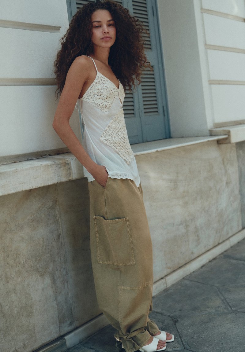 Frau mit lockigem Haar, die ein weißes Spitzen-Camisole und lockere beige Hose trägt, steht mit Händen in den Taschen an einer Marmormauer nahe grauer Fensterläden.