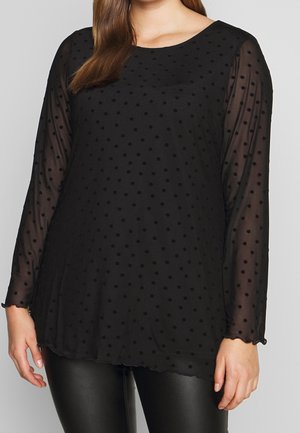 Femme portant un chemisier noir transparent à manches longues à pois, associé à un pantalon en cuir noir, se tenant devant un fond gris.