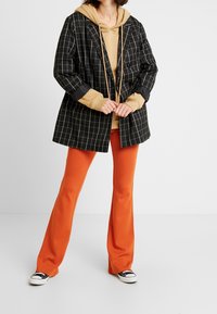 Blazer croisé noir et blanc à carreaux sur un hoodie beige, associé à un pantalon orange évasé et des baskets noires. Textures de tissu lisses.