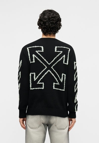 Άτομο με σγουρά σκούρα μαλλιά που φοράει μαύρο πουλόβερ με μεγάλα λευκά σταυρωμένα βέλη στην πλάτη και τα μανίκια, συνδυασμένο με ανοιχτό γκρι τζιν.