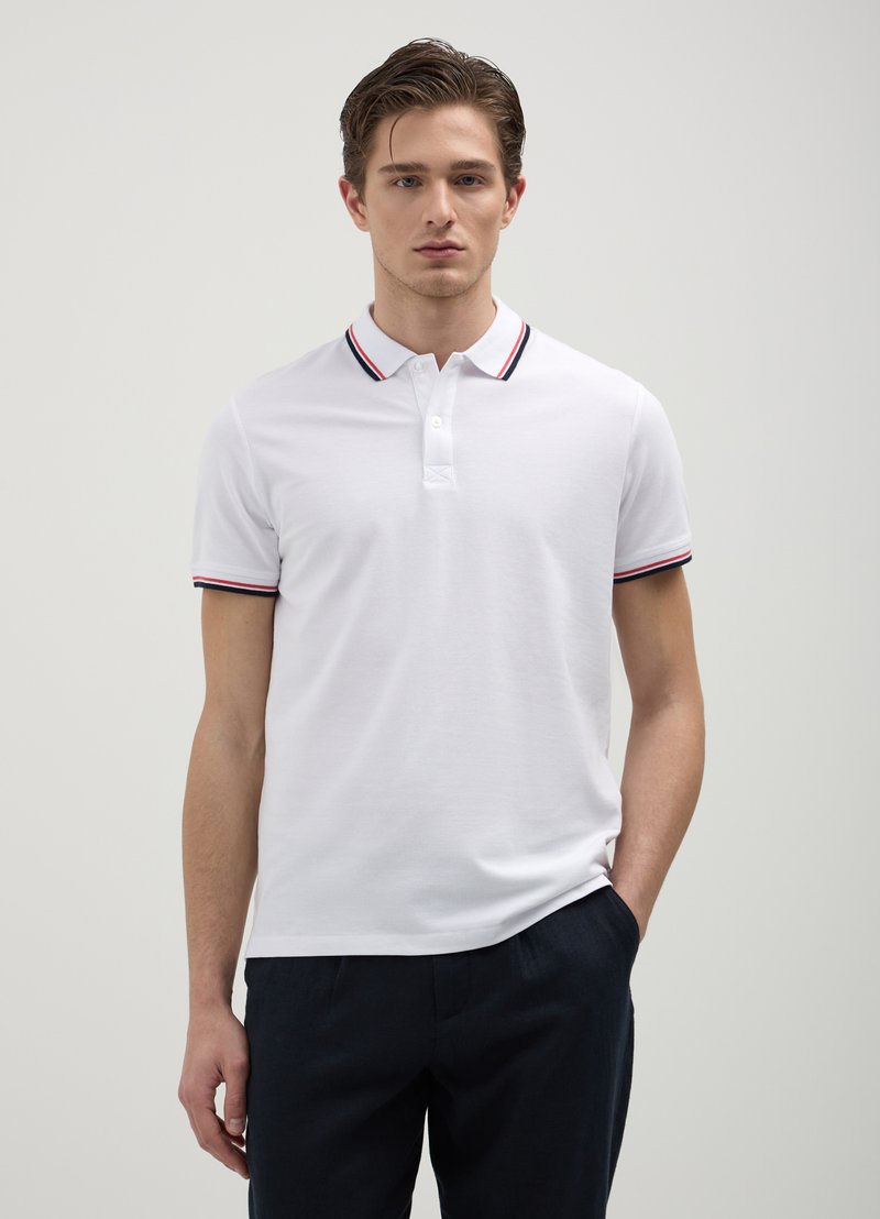Polo bianco con collo a tre bottoni, caratterizzato da un colletto e dei polsini a strisce rosse e blu navy. Indossato con pantaloni blu navy scuri.