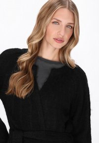 Czarny dziany kardigan o teksturowanym wykończeniu, z paskiem, otwartym przodem i szerokimi rękawami, noszony na ciemnoszarym topie.