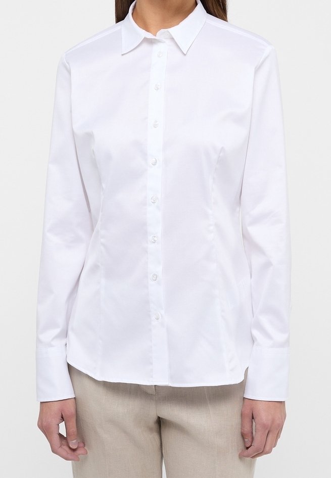 Chemise blanche à manches longues avec col et boutons portée par une personne, associée à un pantalon beige clair sur un fond uni.