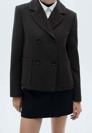 Vrouw draagt een donkerbruine dubbelgeknoopte blazer met grote voorzakken over een wit overhemd met kraag en een zwarte minirok.