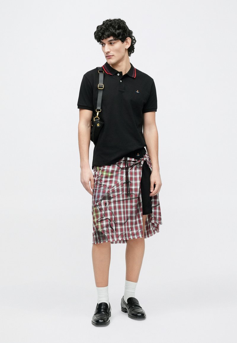 Ung mand iført sort polo, ternet nederdel med sorte shorts under, sorte loafers, hvide sokker og en sort skuldertaske, stående på hvid baggrund.