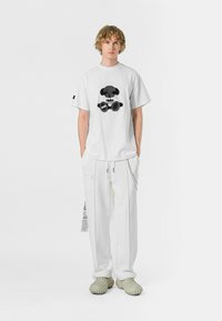 Camisa de algodón blanca con un diseño gráfico negro en la parte delantera, combinada con pantalones blancos holgados y calzado beige texturizado.