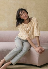 Haut beige texturé avec un motif floral, jeans délavés clairs, assis sur un canapé incurvé rose pâle contre un fond neutre.
