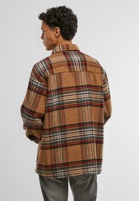 Bruine geruite jas met een wolachtige textuur, voorzien van een kraag, oversized vorm en geweven patronen in beige, zwart en rood.
