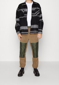 Black and gray checkered jacket with a zip front, paired with beige pants featuring green nylon panels and black slip-on shoes.