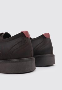 Zapatos de cuero marrón con un acabado suave y labios de talón rojos contrastantes; cuentan con una gruesa suela de goma gris y costuras curvas.