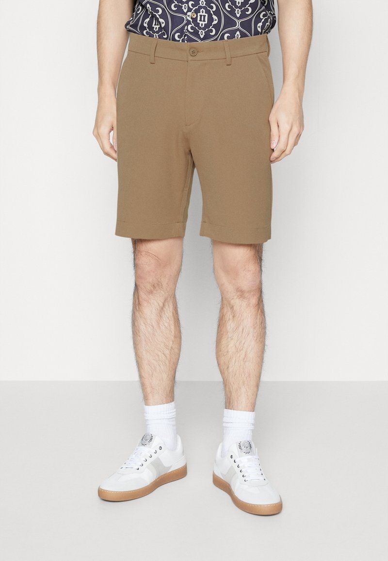 Homme portant un short marron sur mesure, des chaussettes blanches et des baskets blanches à semelles en gomme, debout devant un fond uni.