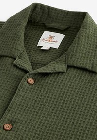 Chemise en maille texturée vert olive avec boutons en bois et col, avec une étiquette blanche comportant un texte et un logo de palmier.