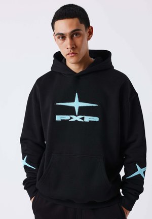 Jeune homme portant un sweat à capuche noir avec une étoile bleu clair et le texte "PXP", mains dans les poches, expression neutre, fond clair uni.