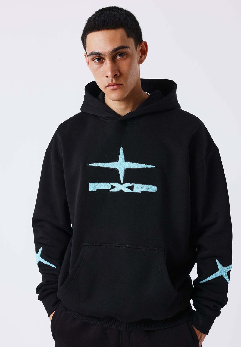 Jeune homme portant un sweat à capuche noir avec une étoile bleu clair et le texte "PXP", mains dans les poches, expression neutre, fond clair uni.