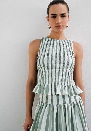 Femeie purtând o bluză fără mâneci, cu dungi verticale verzi și albe, și o fustă asortată cu talie strânsă și tiv peplum, stând în fața unui perete alb.