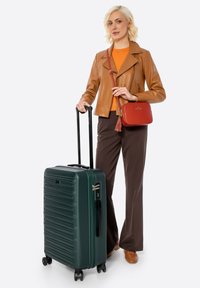 Blondýnka v hnědé kožené bundě, oranžovém topu a hnědých kalhotách, která drží tmavě zelený kufr na kolečkách a červenou crossbody tašku.