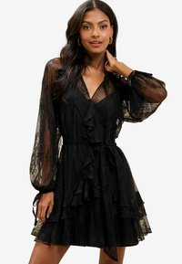 Schwarzes Spitzenkleid mit langen transparenten Ärmeln, Rüschendetails und einem Bindegürtel. Besonderheiten sind der geschichtete Rock und das V-Ausschnitt-Design.